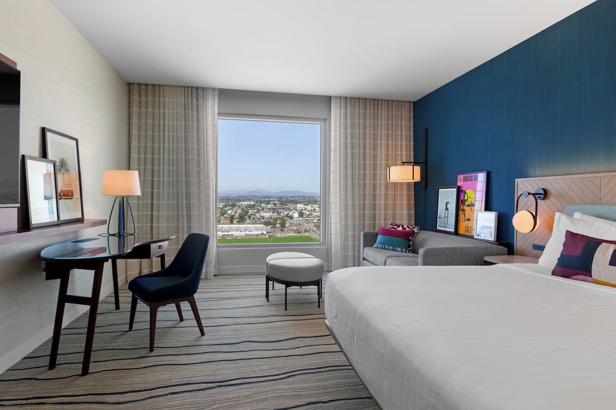 Hotelkamer met een groot bed, een bureau met stoel, een klein tafeltje en uitzicht op een stadsbeeld door een groot raam.
