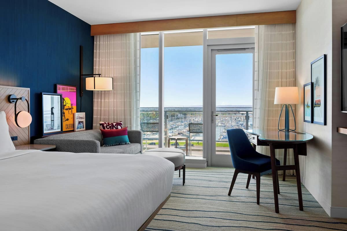 Een hotelkamer met een groot bed, een bureau, een stoel en uitzicht op de stad door grote ramen.