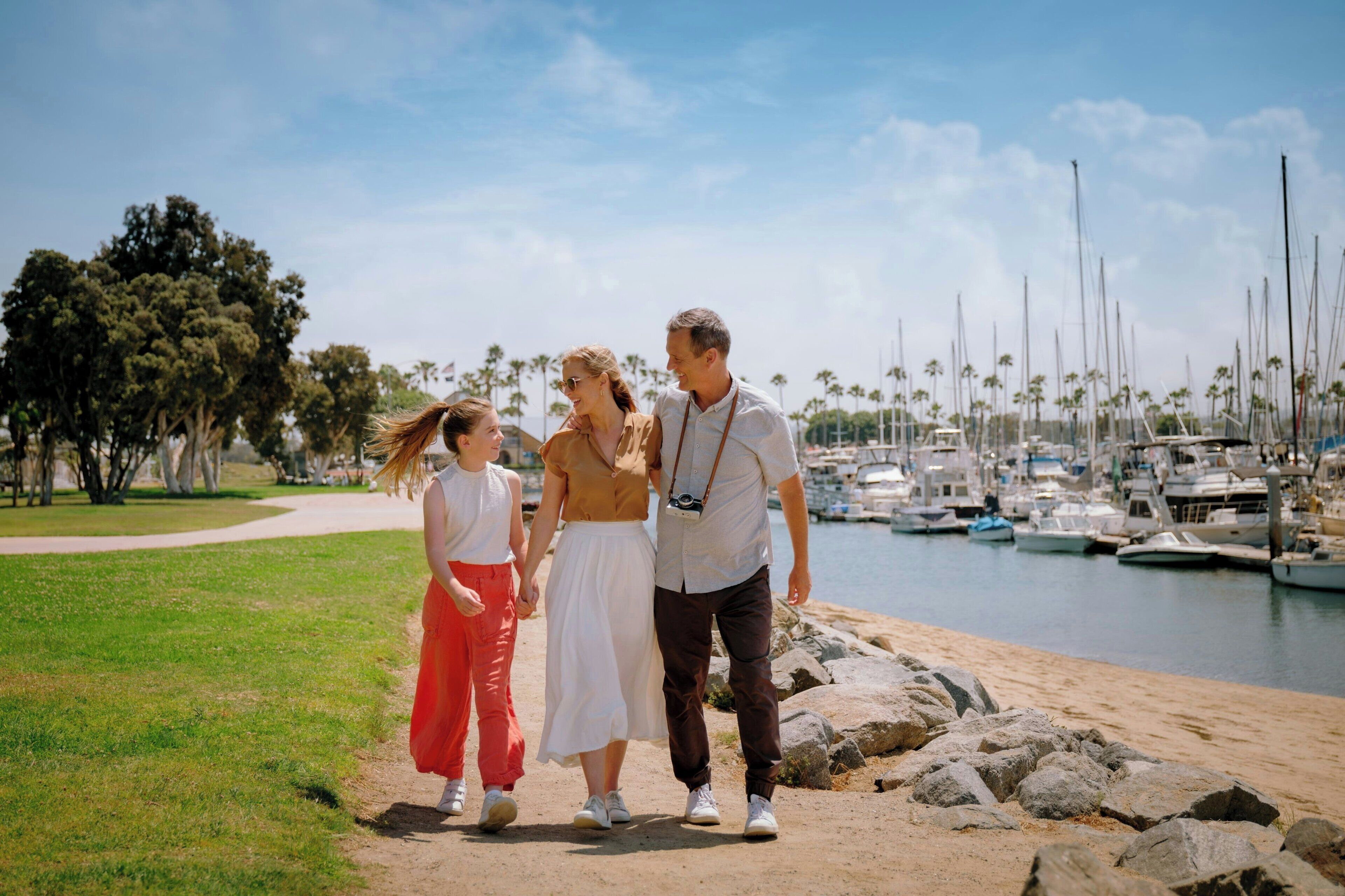 A family walking along a sandy path by a marina with boats in the background.