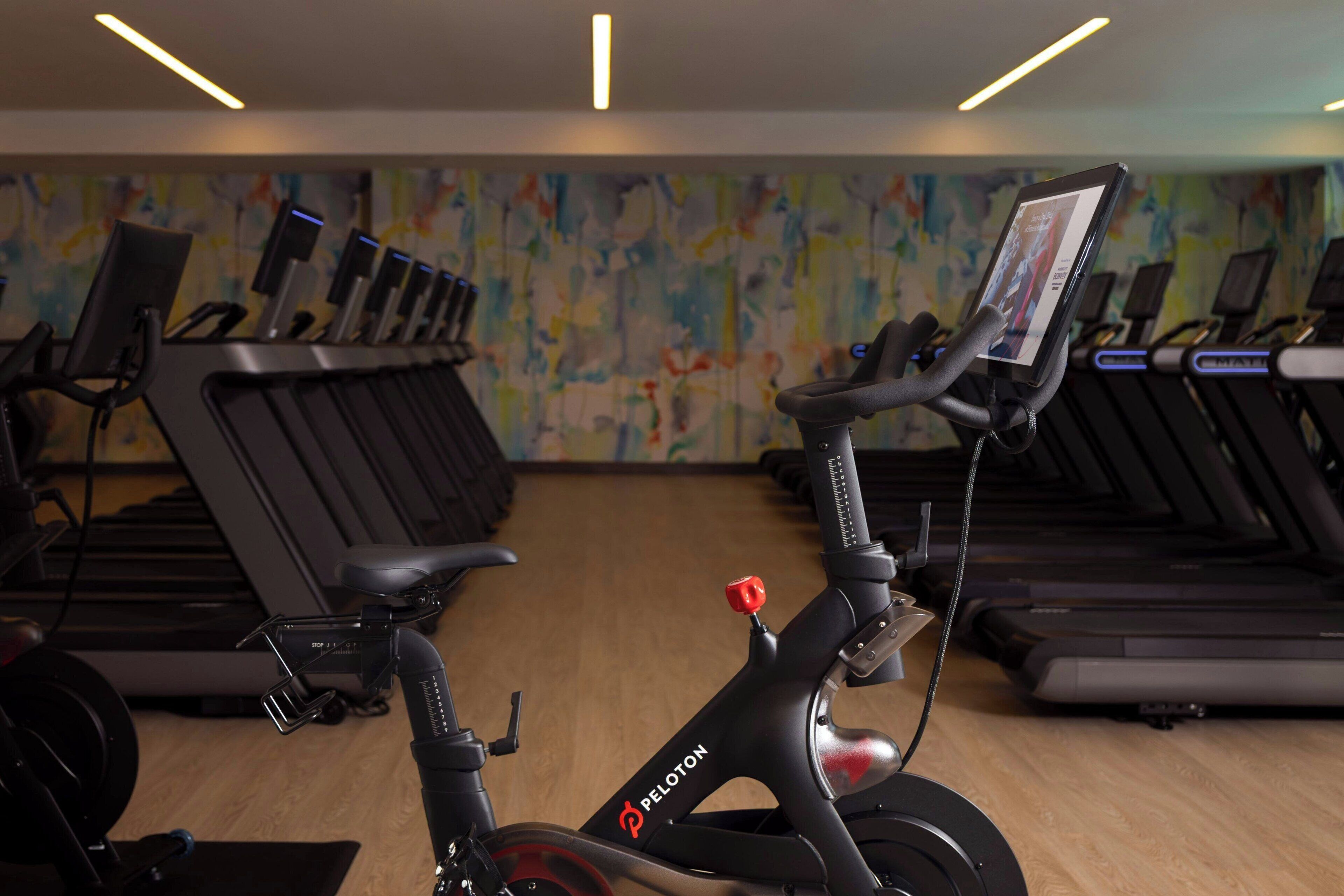 A gym with stationary bikes and treadmills, a colorful abstract wall, and a wooden floor.