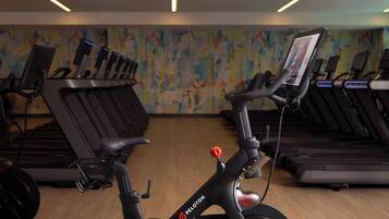 A gym with stationary bikes and treadmills, a colorful abstract wall, and a wooden floor.