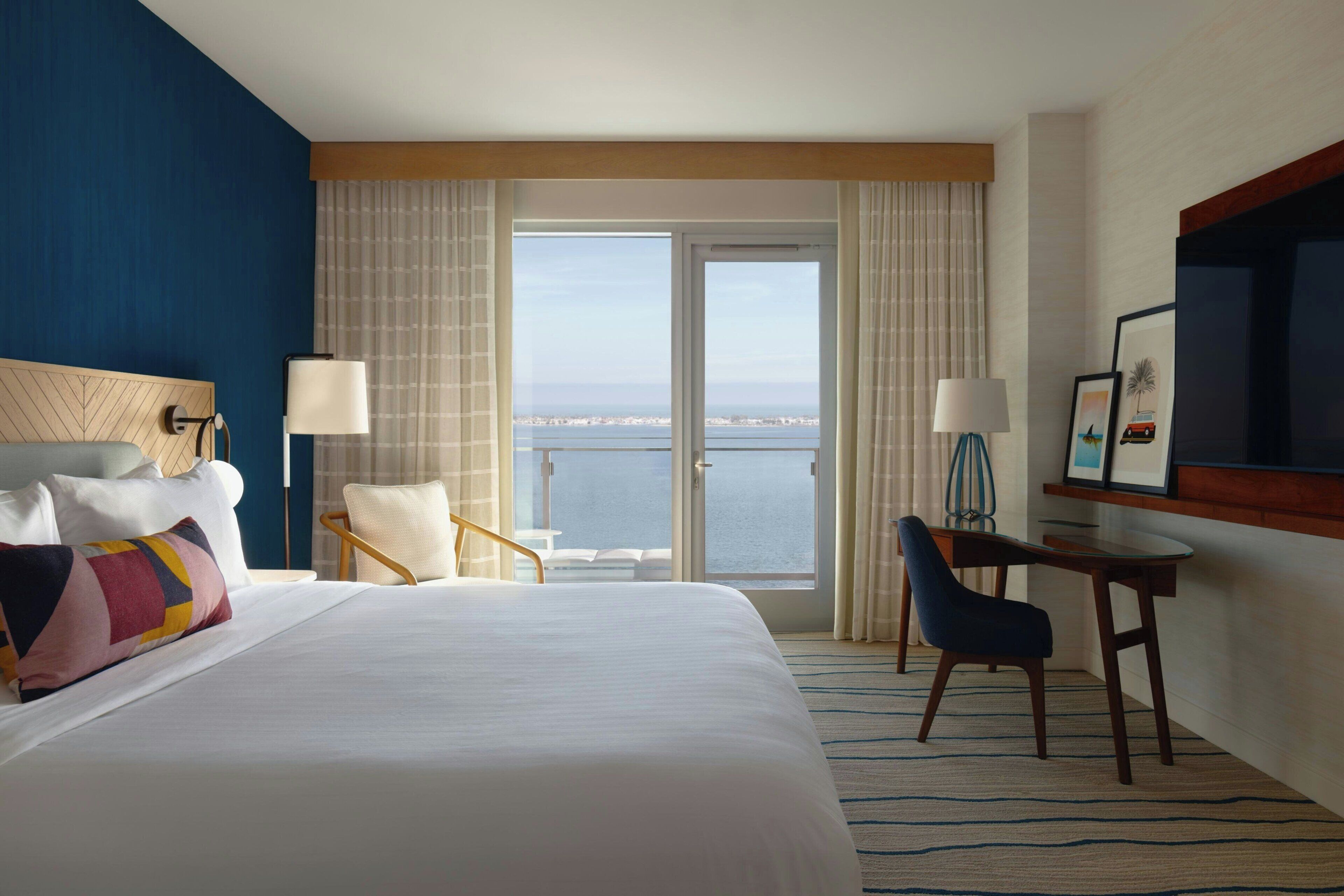 A hotel room with a large bed, a desk, a chair, and a view of the sea through a sliding door.