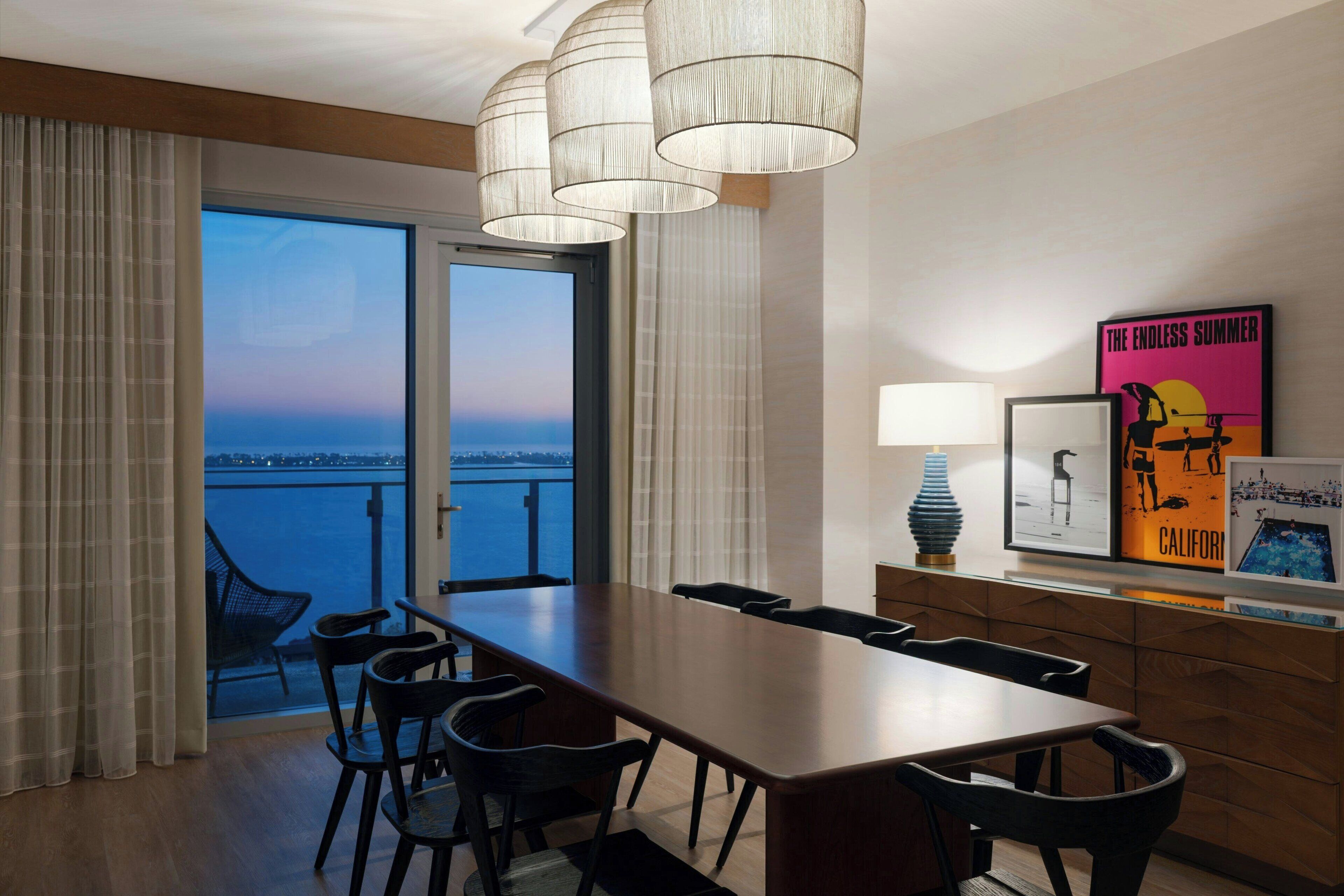 Habitación de hotel moderna con una gran mesa de comedor, sillas, vistas de la ciudad y un balcón con cortinas.