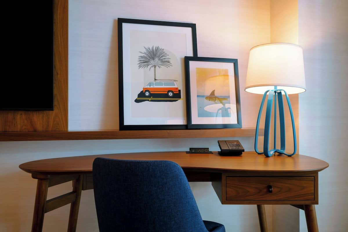 Een hotelkamer met een houten bureau, een blauwe stoel, een lamp en twee ingelijste foto's aan de muur.