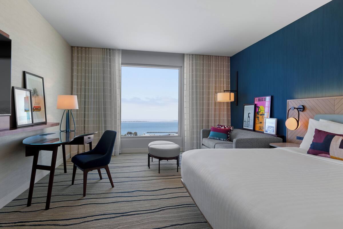 Een hotelkamer met een groot bed, een bureau met stoel, een klein voetenbankje en uitzicht op de oceaan.