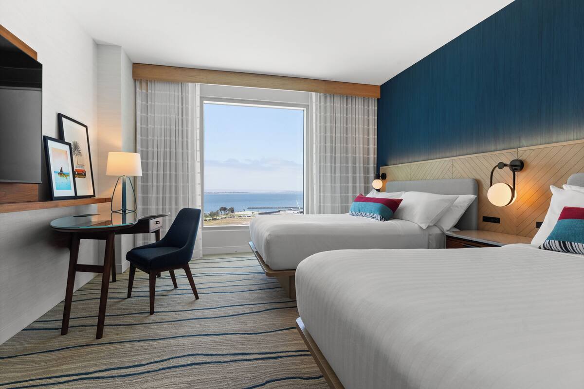 Hotelkamer met twee bedden, een bureau, een stoel, een televisie en een groot raam met uitzicht op de oceaan.