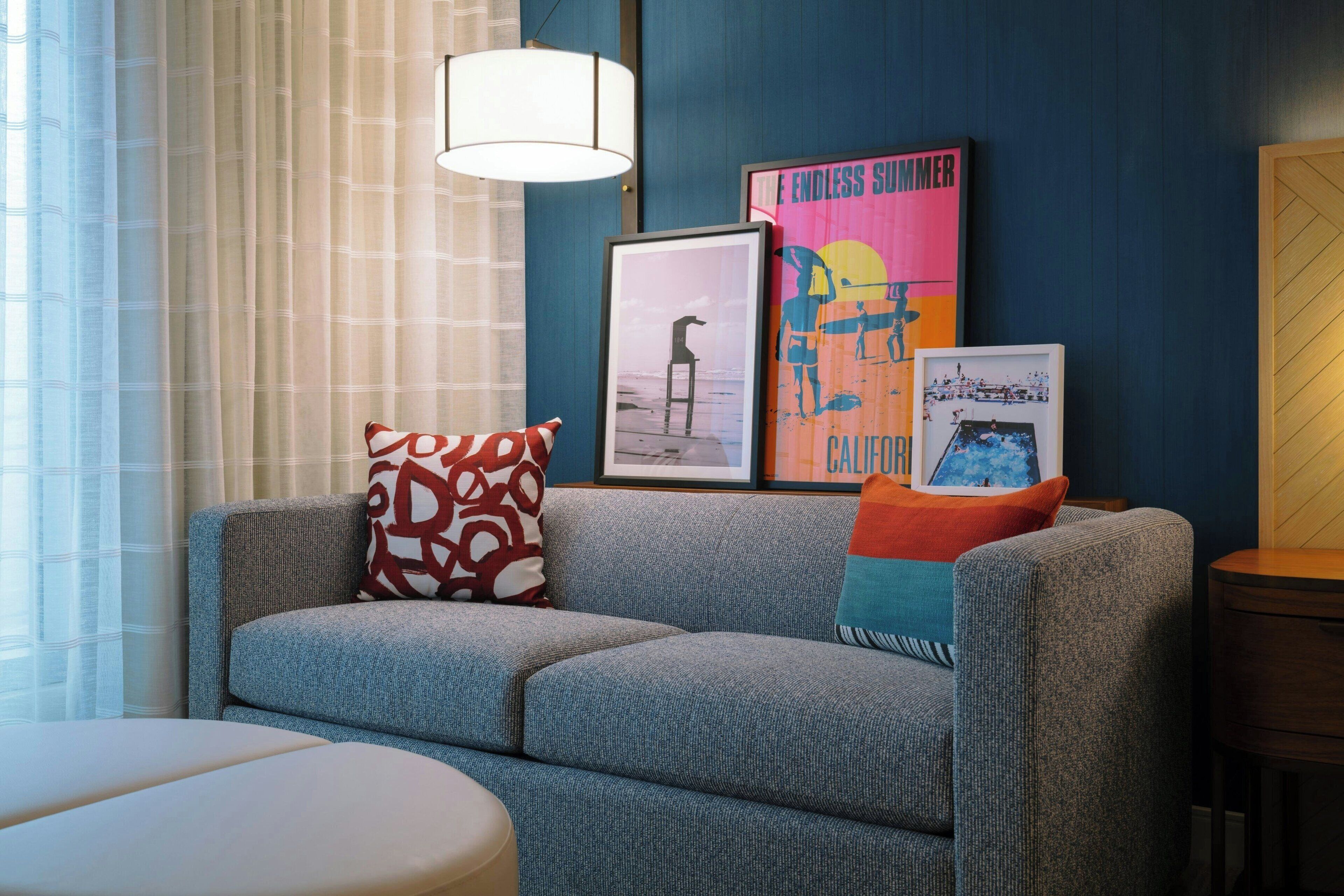A modern living room with a grey sofa, a white ottoman, and a wooden side table. There are three framed pictures on the wall, one of which is titled "Endless Summer."