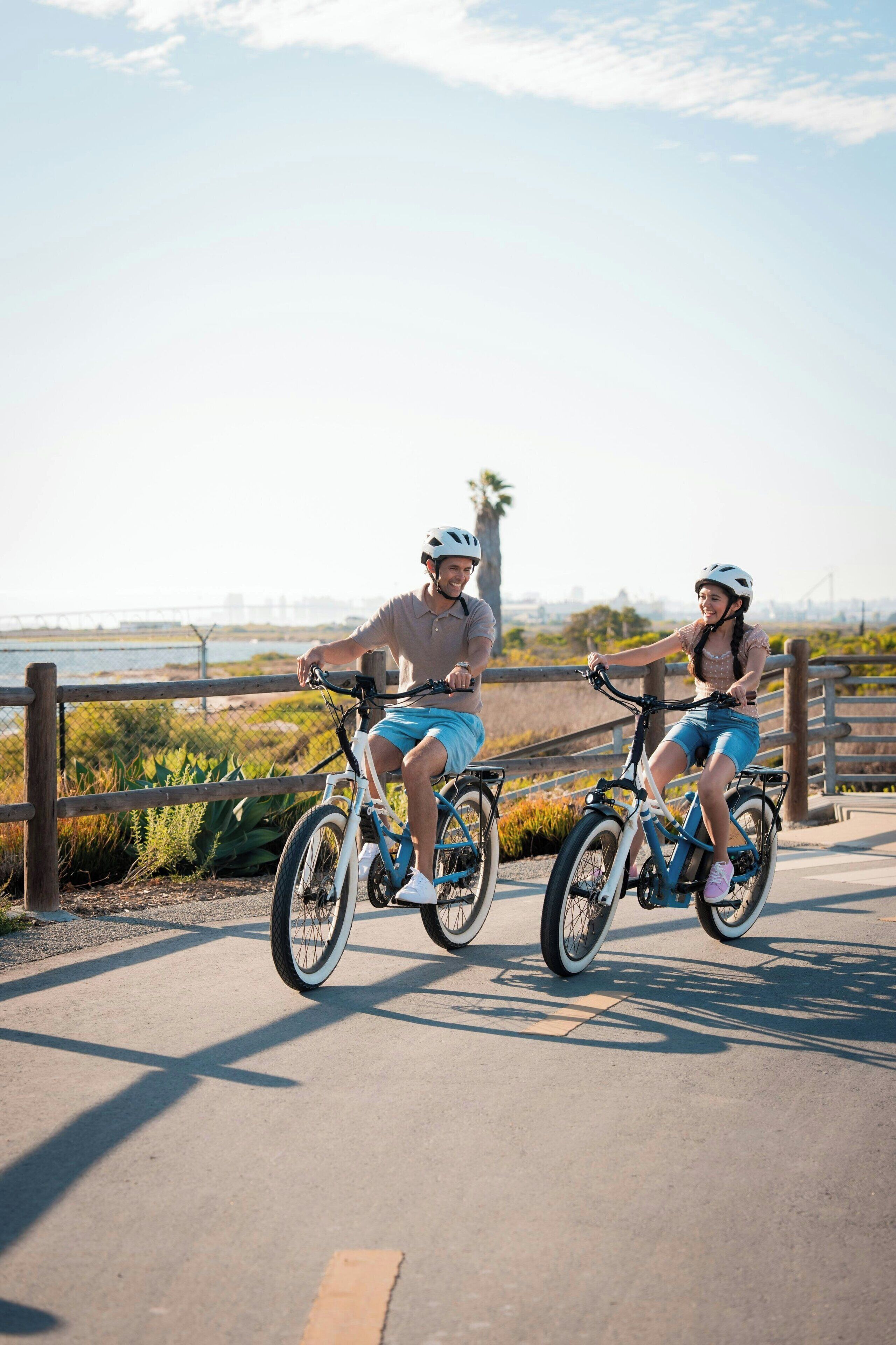 Two people riding bicycles on a paved path with a wooden fence and greenery in the background.