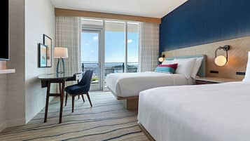 A hotel room with two beds, a desk with a chair, a TV, and a view of the city through large windows.