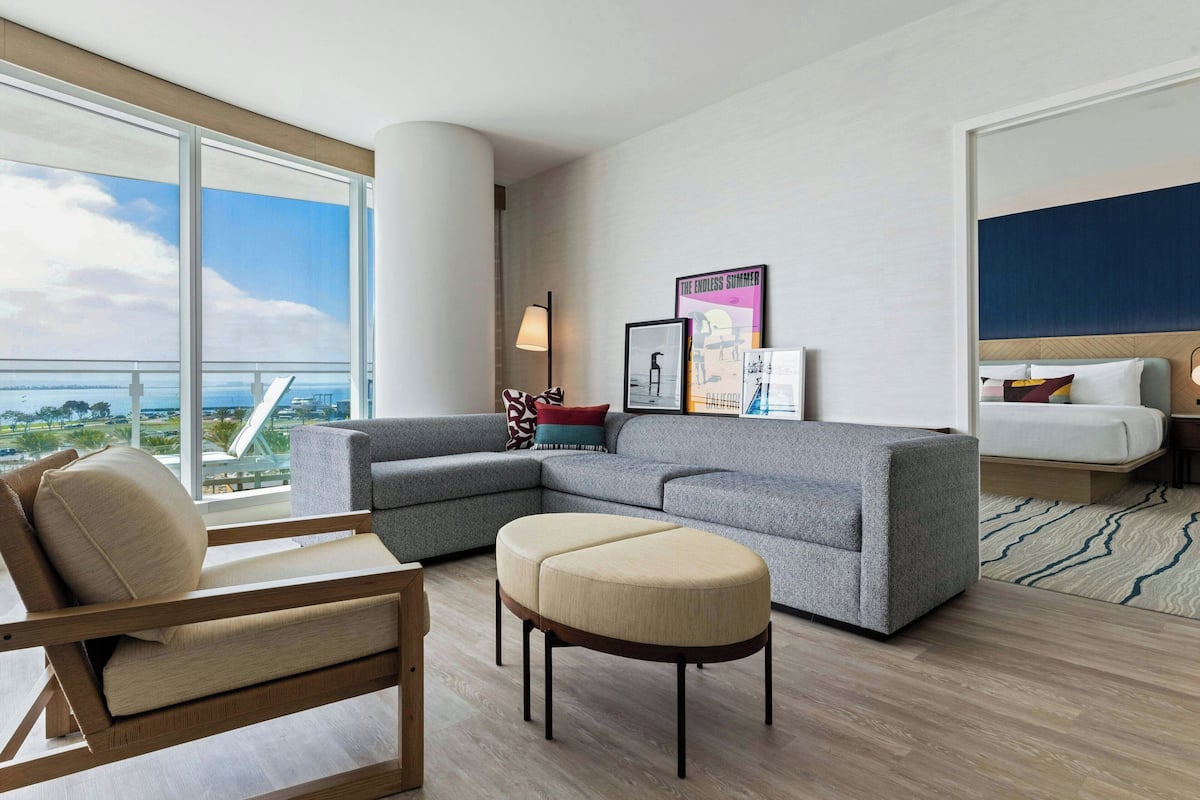 Een moderne hotelkamer met een grijze modulaire bank, een houten fauteuil, een beige voetenbankje en een bed met een blauwe hoofdbord. Er hangen ingelijste foto's aan de muur en er is uitzicht op het landschap door grote ramen.