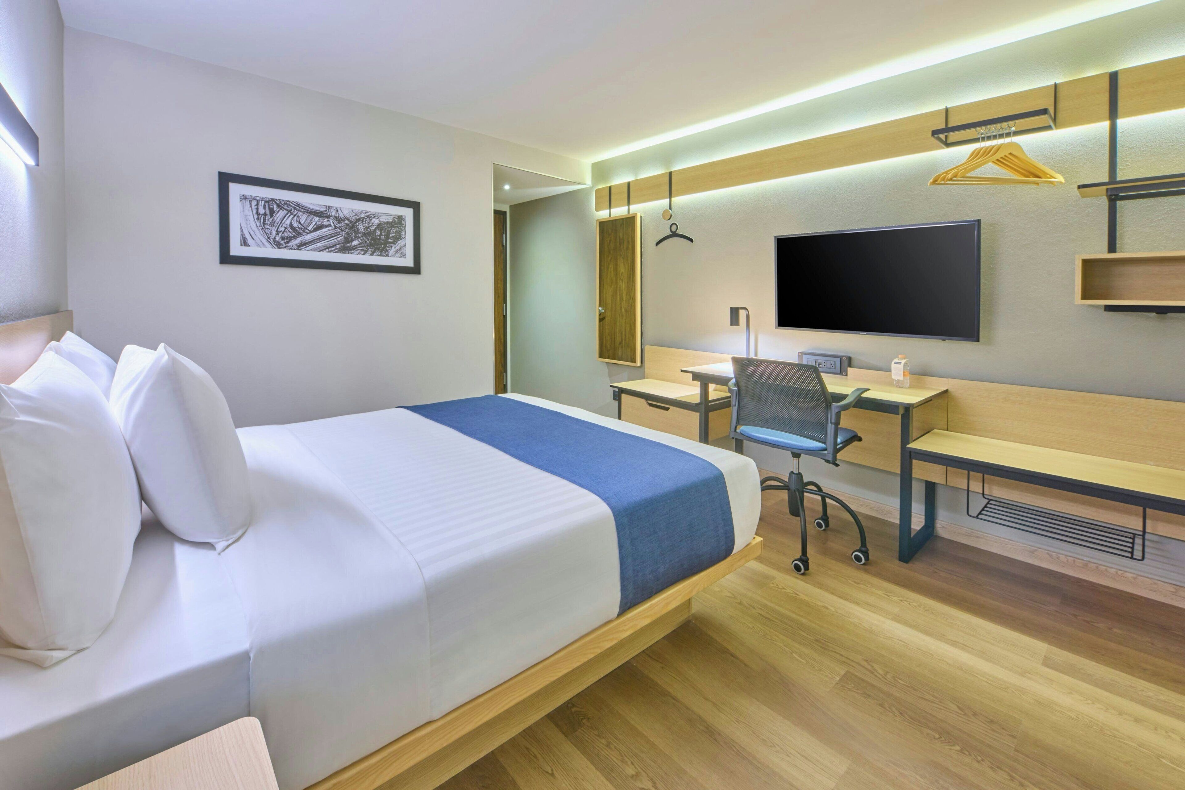 A hotel room with a bed, desk, chair, TV, and a framed picture on the wall.