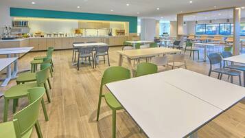 A modern dining area with multiple tables and chairs, a kitchen in the background, and large windows providing natural light.
