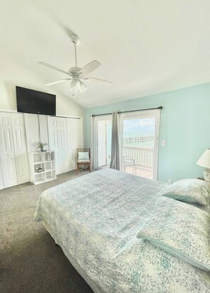 Un dormitorio con una cama grande, un ventilador de techo, un televisor, un armario, una pequeña zona de estar y un balcón con vista al mar.