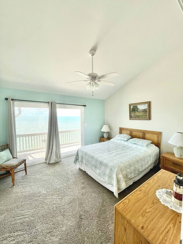 A bedroom with a bed, a wooden nightstand, a desk, a chair, a ceiling fan, and a view of the ocean through large windows.