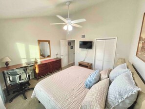 A bedroom with a bed, dresser, desk, chair, ceiling fan, TV, and closet.