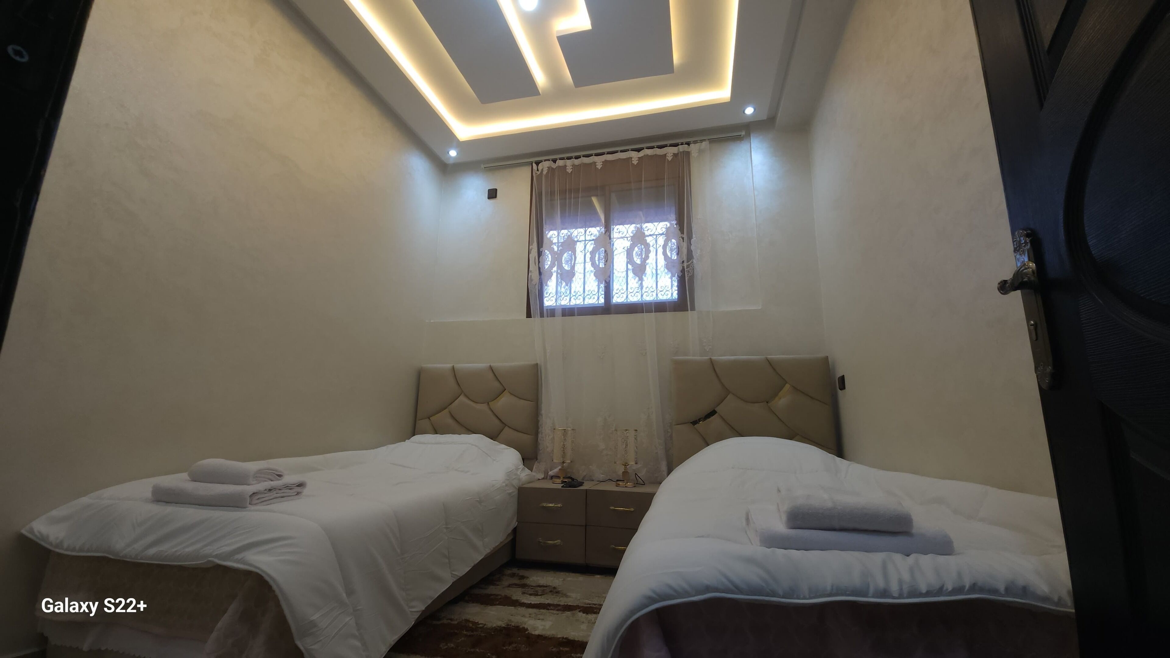 Ein Schlafzimmer mit zwei Betten, einem Fenster mit dekorativen Vorhängen und einer Decke mit Einbauleuchten.