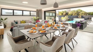 A modern dining area with a table set for breakfast, surrounded by comfortable seating and large windows offering a view of a pool and greenery. - ODIN 2 by Buenavilla | Experience Unmatched Luxury in Puerto Calero (Puerto Calero)