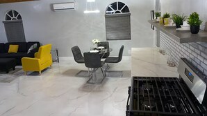Eine moderne Küche mit Gasherd, ein Essbereich mit Glastisch und Stühlen sowie ein Wohnbereich mit einer Couch und einem gelben Sessel.