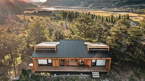 Una casa moderna con techo plano, ventanas amplias y una terraza de madera, rodeada de bosque y montañas.