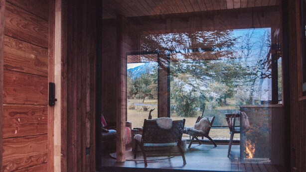 Interior de una cabaña de madera con un ventanal que da a una zona exterior con sillas y una chimenea.
