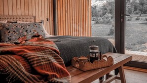 Un dormitorio con cabecera de madera, cama con ropa de cama estampada con motivos florales y cuadros, mesita de noche con un farol y cestas, y vistas a un paisaje nevado a través de un amplio ventanal.