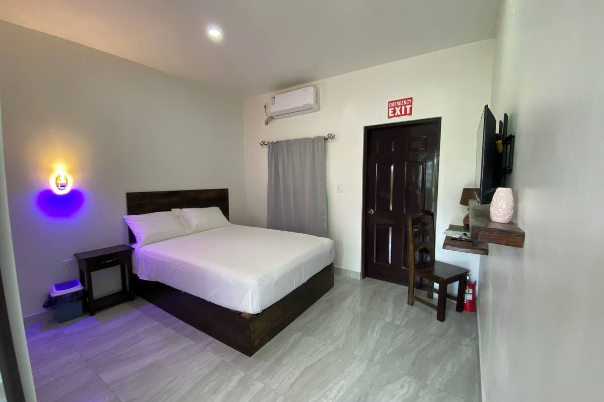 A hotel room with a bed, bedside table, nightstand, and a wall-mounted air conditioner.