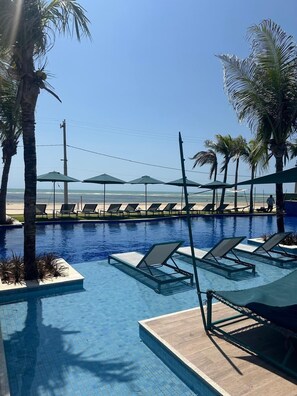 A pool area with lounge chairs and umbrellas, palm trees, and a view of the ocean. - Casa Flecheiras Condomínio Azure (Trairi)