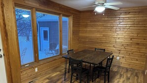 Un comedor con mesa y sillas de madera, ventanas grandes con persianas y un ventilador de techo.