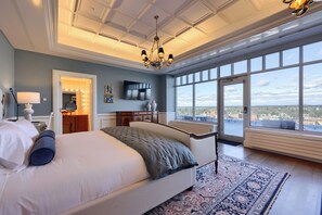 A spacious bedroom with a large bed, a chandelier, a large window with a city view, and a walk-in closet. - The Executive Suites and Venue (Ephrata)