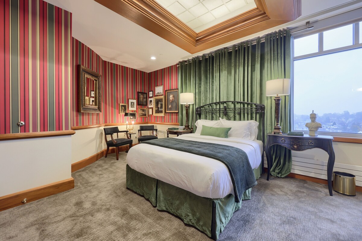 A hotel room with a large bed, a desk with a lamp, a chair, and a view of the city through a window.