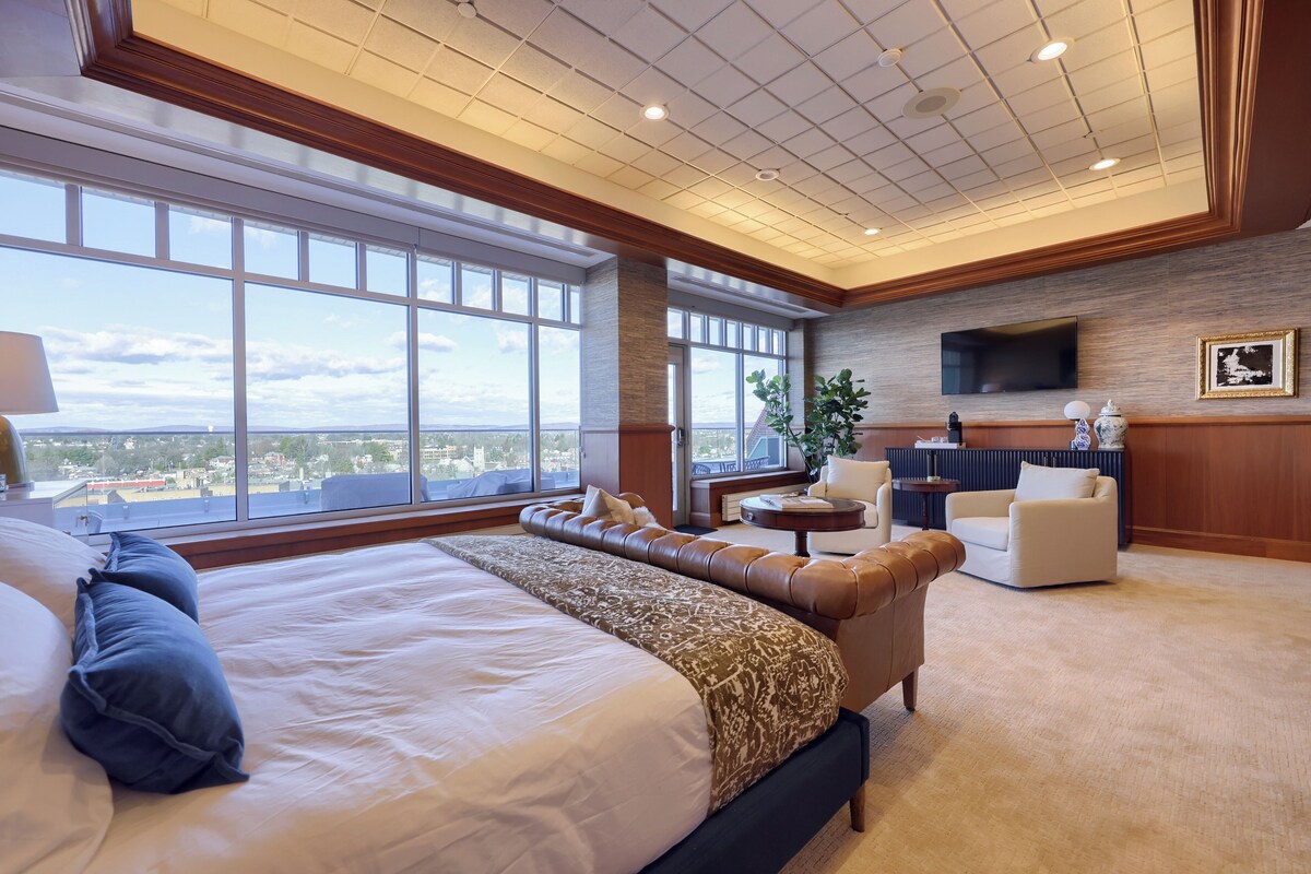 A spacious bedroom with a large bed, a seating area with a sofa and armchairs, a television mounted on the wall, and a large window offering a city view.
