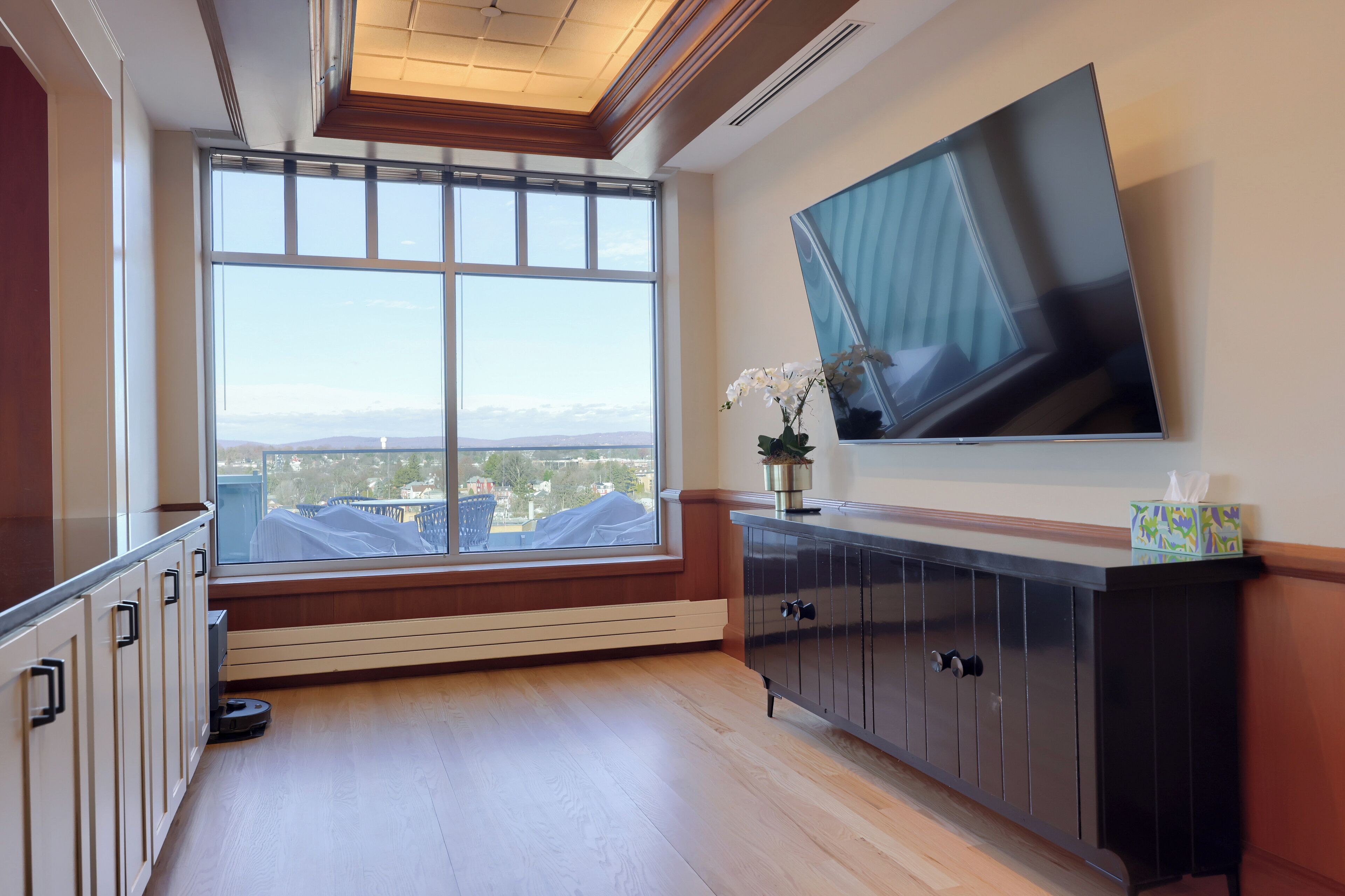 A modern room with a large window, a flat-screen TV mounted on the wall, and a dark wooden cabinet.