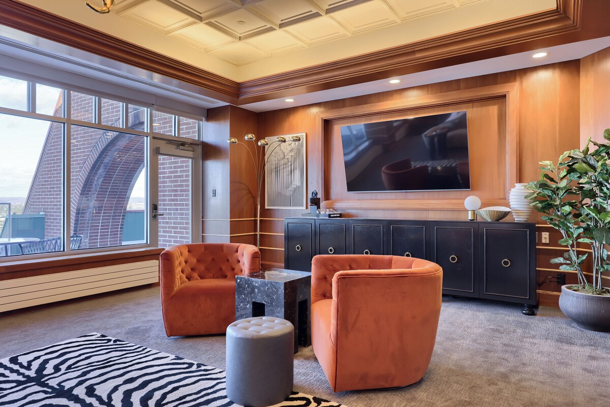 A modern living room with orange armchairs, a black entertainment unit, and a large window with a city view.