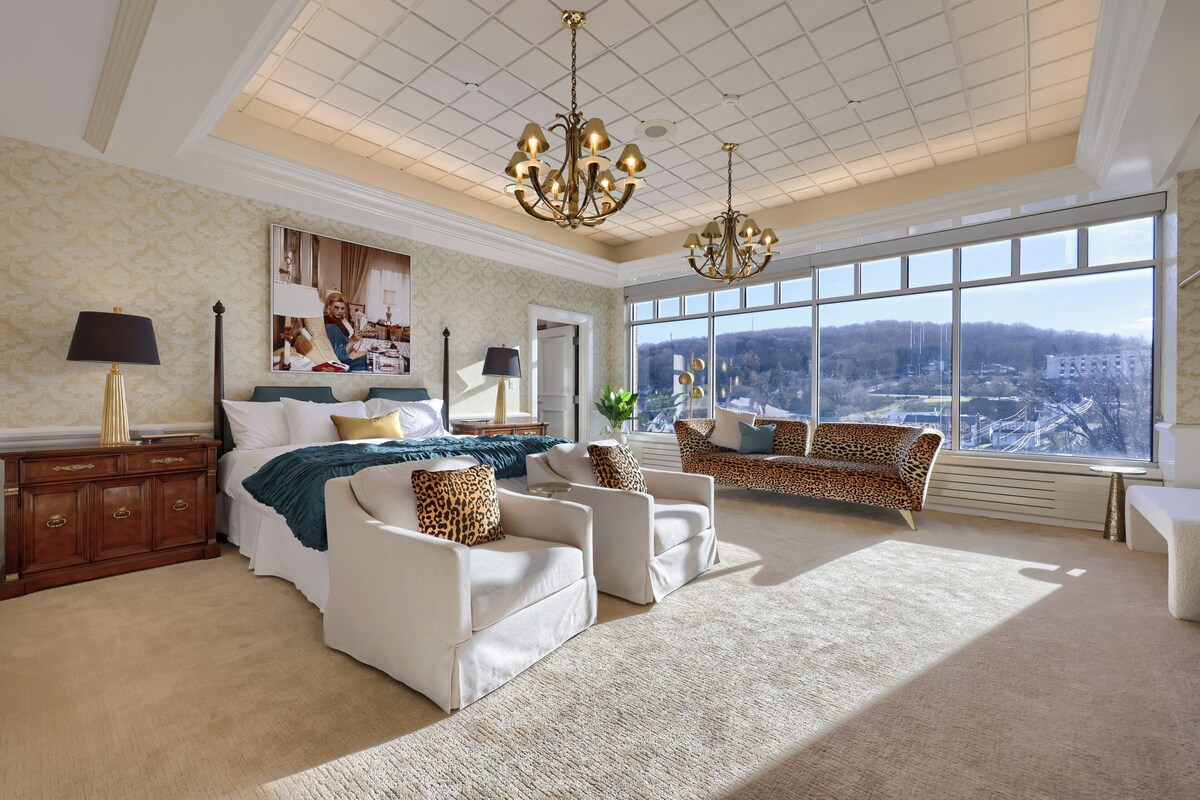 A spacious bedroom with a large bed, two sofas, a chaise lounge, and a view of a cityscape through large windows.