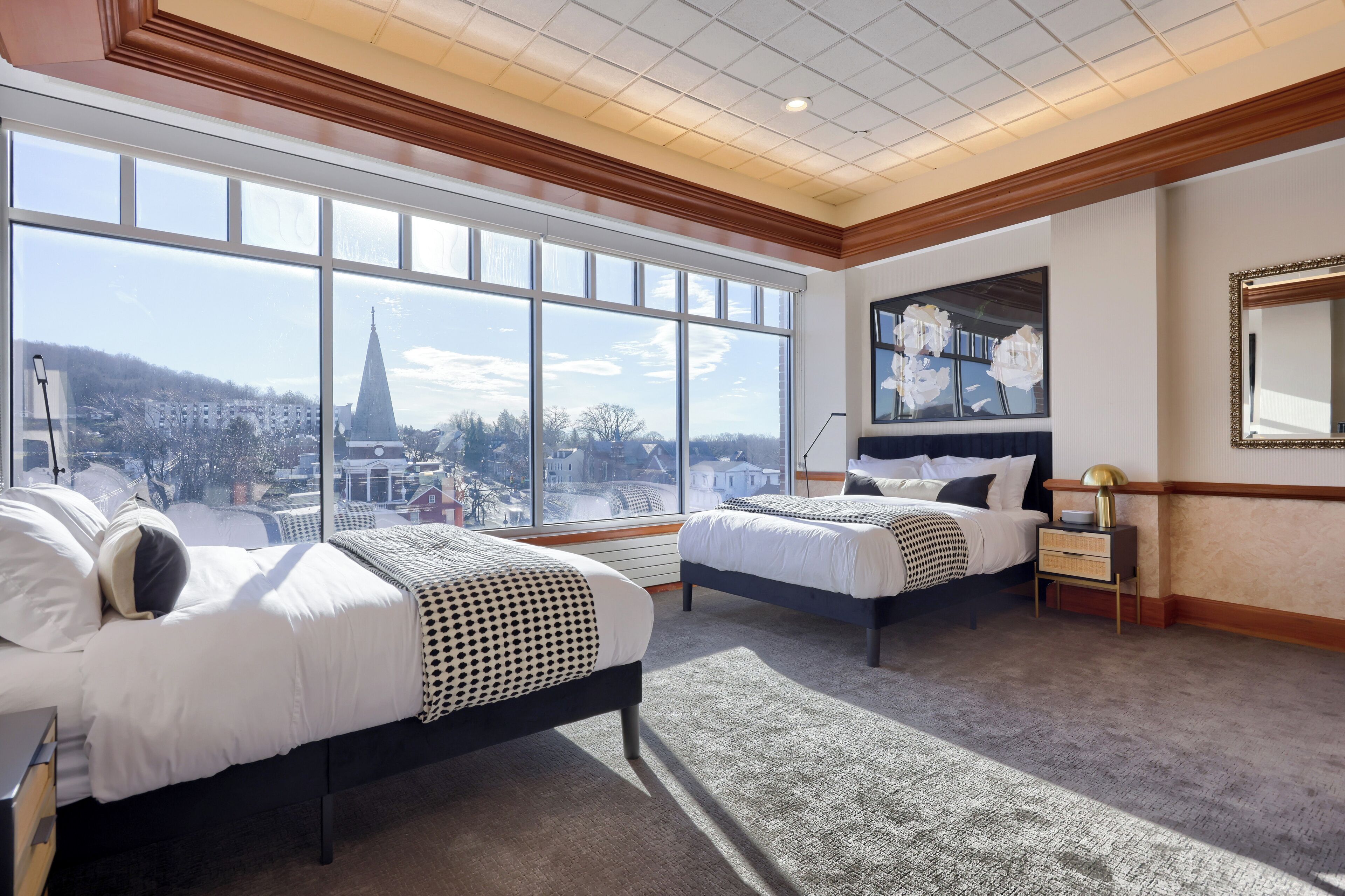 A hotel room with two beds, large windows, a city view, a painting, and a mirror.