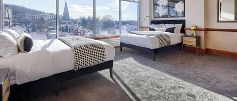 A hotel room with two beds, large windows, a city view, a painting, and a mirror.