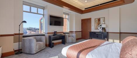A hotel room with a large bed, a desk with a chair, a television, and a window with a view of a building.