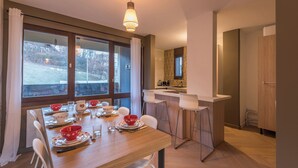 Un coin repas avec une table en bois dressée pour quatre personnes, une cuisine en arrière-plan et une grande fenêtre avec des rideaux.
