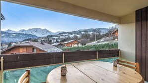 Un balcon avec une table et des chaises en bois, une clôture en bois et une vue sur des montagnes et des maisons enneigées.