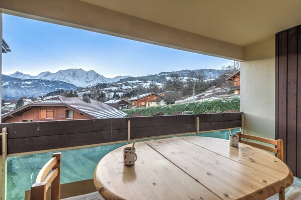 A balcony with a wooden table and chairs, a wooden fence, and a view of snow-covered mountains and houses. - Appartement Alpestre - Welkeys (Combloux)