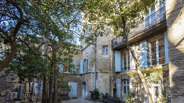 Ein Innenhof mit Kiesweg, Bäumen und einem Gebäude mit Steinmauern und Fensterläden.