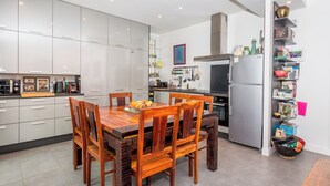 A modern kitchen with white cabinets, a wooden dining table, and chairs. There is a refrigerator, a stove, and various kitchen appliances. - Appartement City Garden - Welkeys (Biarritz)