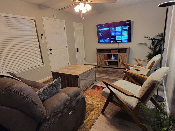 Una sala de estar con un televisor montado en la pared, una mesa de centro de madera, un sofá de cuero marrón y un sillón color beige.