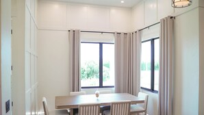 Un comedor con mesa rectangular y seis sillas, ventanas amplias con cortinas, y una lámpara colgante.