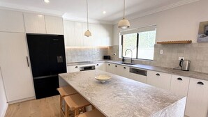 A modern kitchen with a large island, wooden stools, a black refrigerator, and pendant lighting. - Amazing 3bd in National Park (Bundeena)