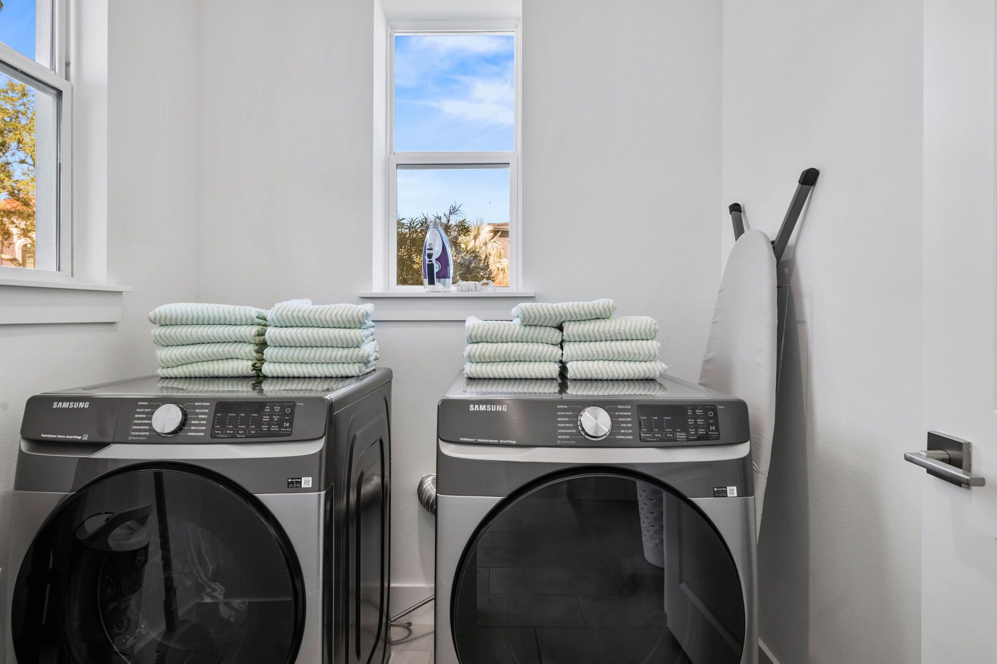 A laundry area with two Samsung washing machines, a window with a view of palm trees, and a drying rack.