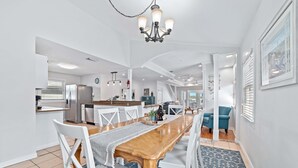 A spacious dining area with a wooden table and white chairs, a chandelier, and a view of the kitchen in the background.