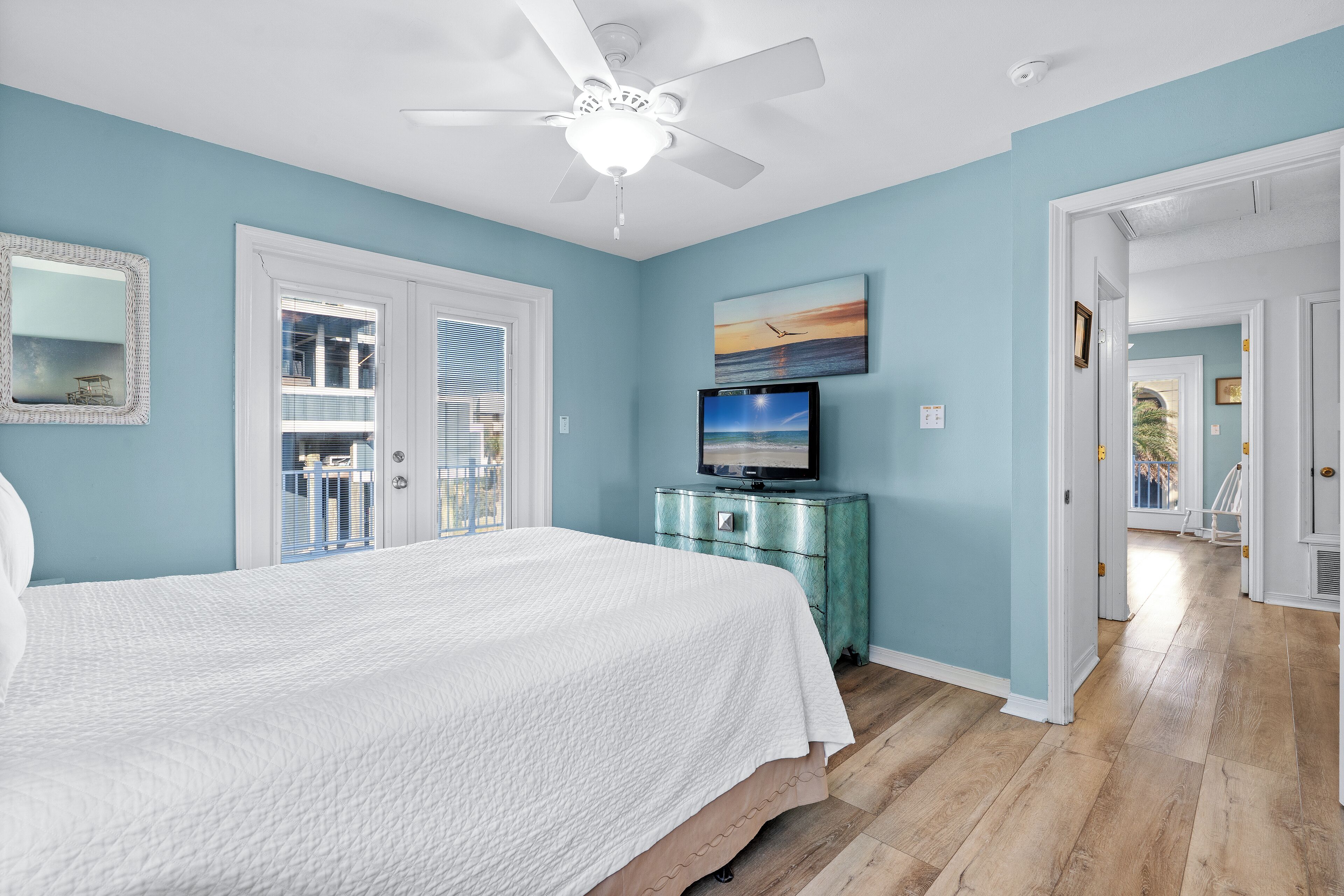 A bedroom with a large bed, a ceiling fan, a TV on a teal dresser, and a view of the outside through a door.
