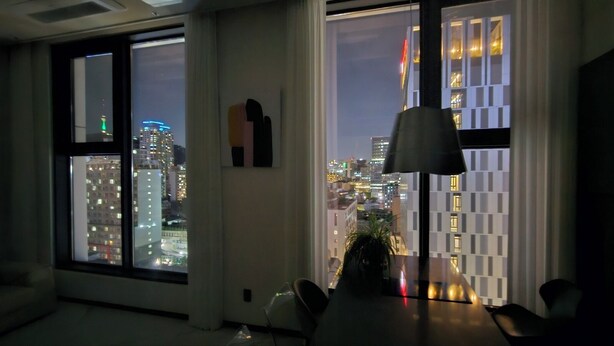 Een kamer met grote ramen en uitzicht op de stad, een moderne lamp en een klein plantje op de vensterbank.