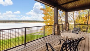 A covered outdoor deck with a table and chairs, overlooking a lake and autumn foliage. - Large Home | Reservoir Views & Access | Hot Tub | Fire Pit | Basketball Court (Island Park)